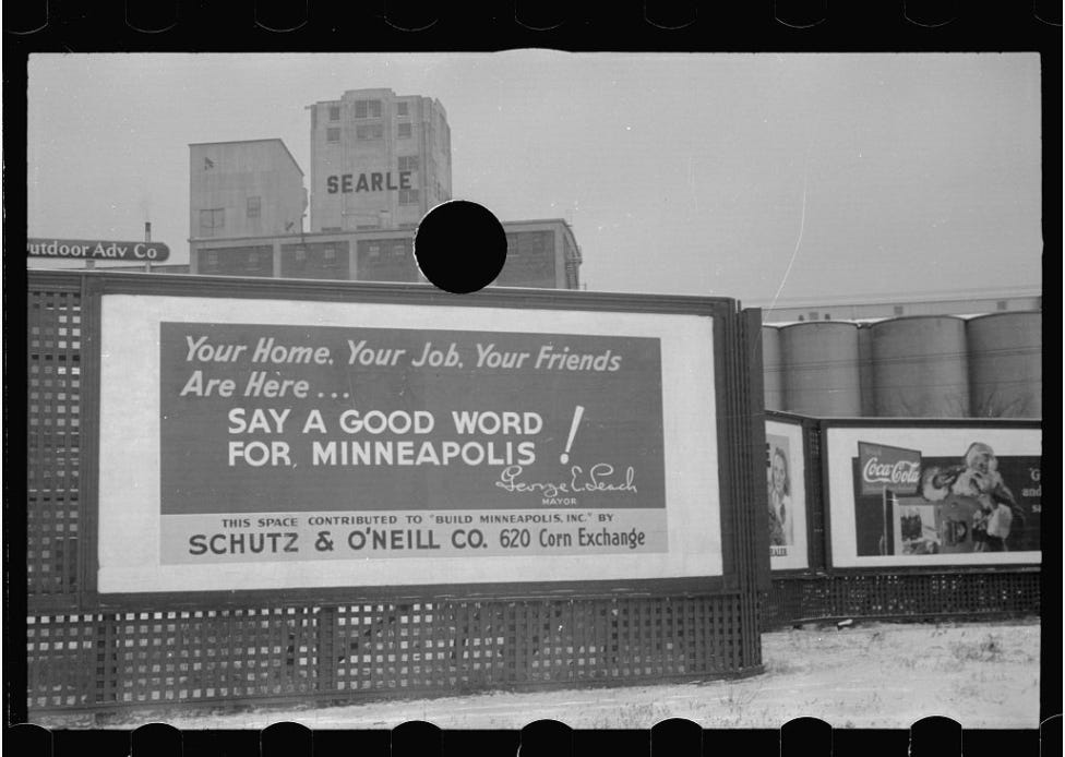 Black and white photo of billboard in front of grain elevators. The billboard reads: "Your home, your job, your friends are here... Say a Good Word For Minneapolis!"
