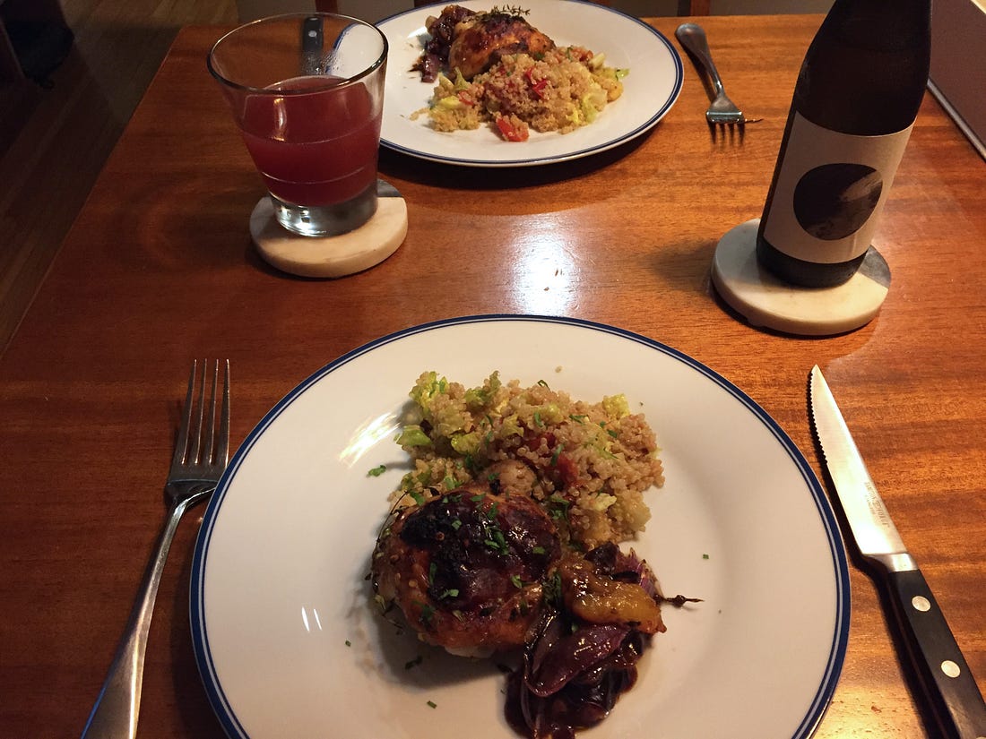 On a polished wood table, two plates with quinoa salad and a chicken thigh with dark, crispy skin. Next to the chicken is a small pile of roasted plums and onions, with a dark sauce pooling at the bottom.