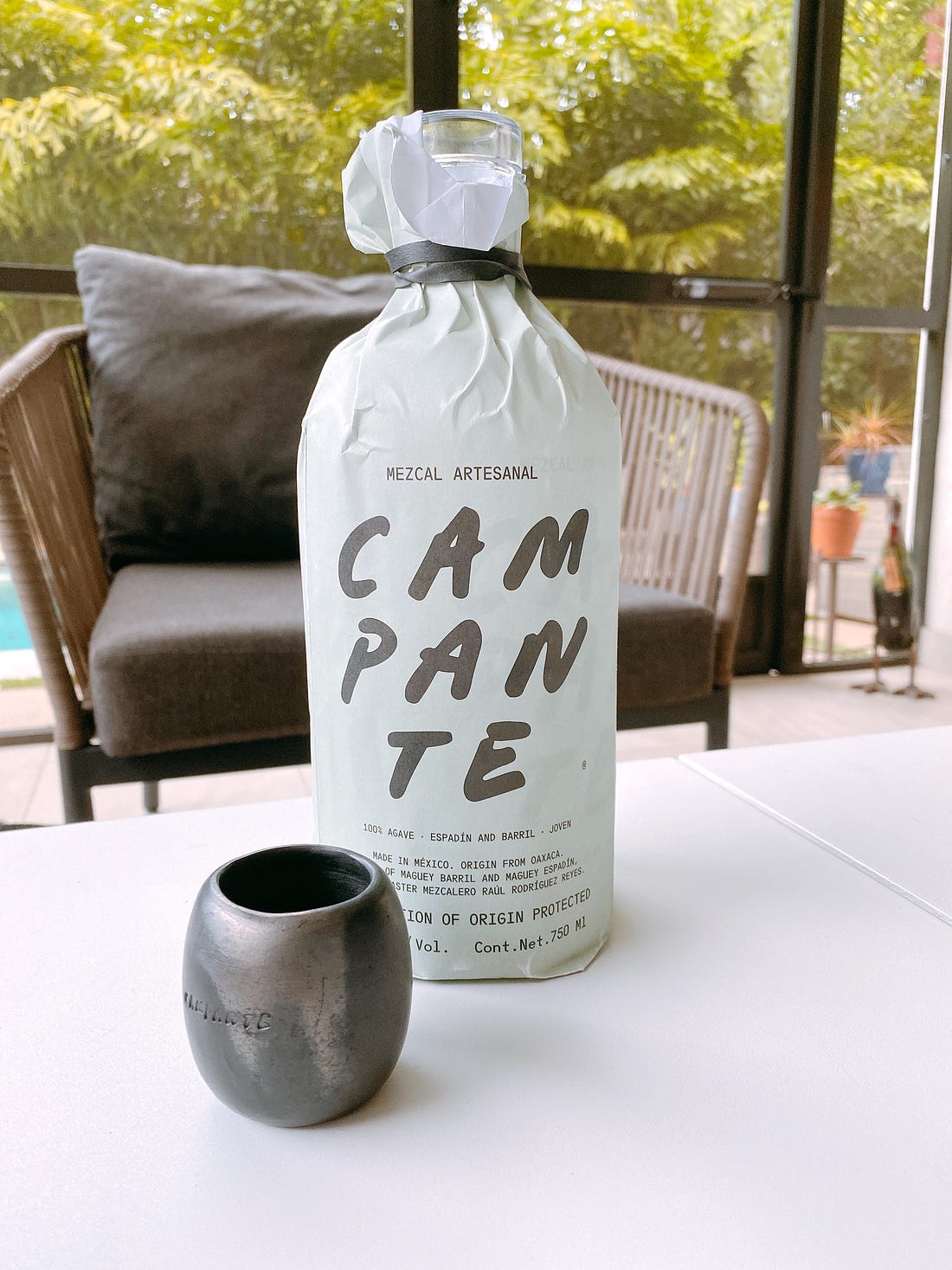 A bottle of Mezcal Campante and a traditional clay cup is in the foreground. In the background, a gray chair sits in front of a screen, behind which there is a pool, potted plants, and lush tropical greenery.