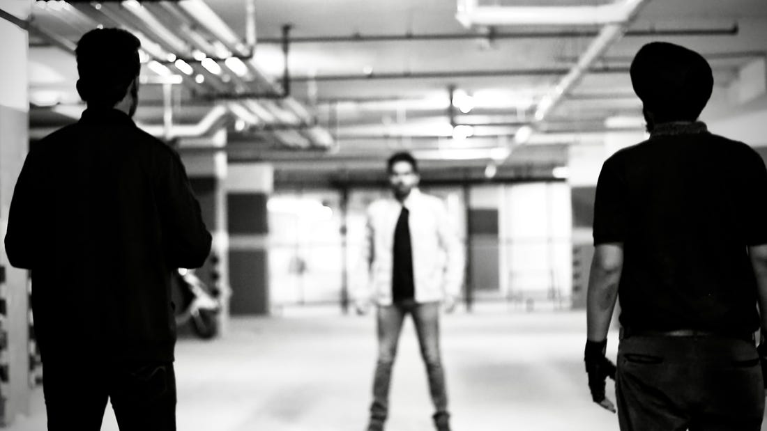 From 'The Glint of Darkness: Revolution': A man in a parking garage, slightly out of focus and facing the camera from far away, is confronted by two men in black shirts, in focus, backs to the camera.