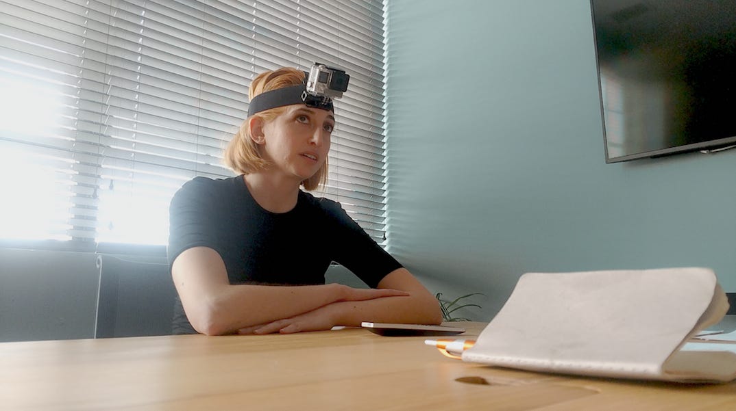 From the film 'I Blame Society': A blonde woman sits at a desk, wearing a Go-Pro camera on her forehead, looking at unseen characters in the room.