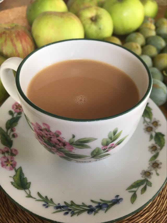 A cup of tea on a saucer with flowers on and apples and greengages in the background