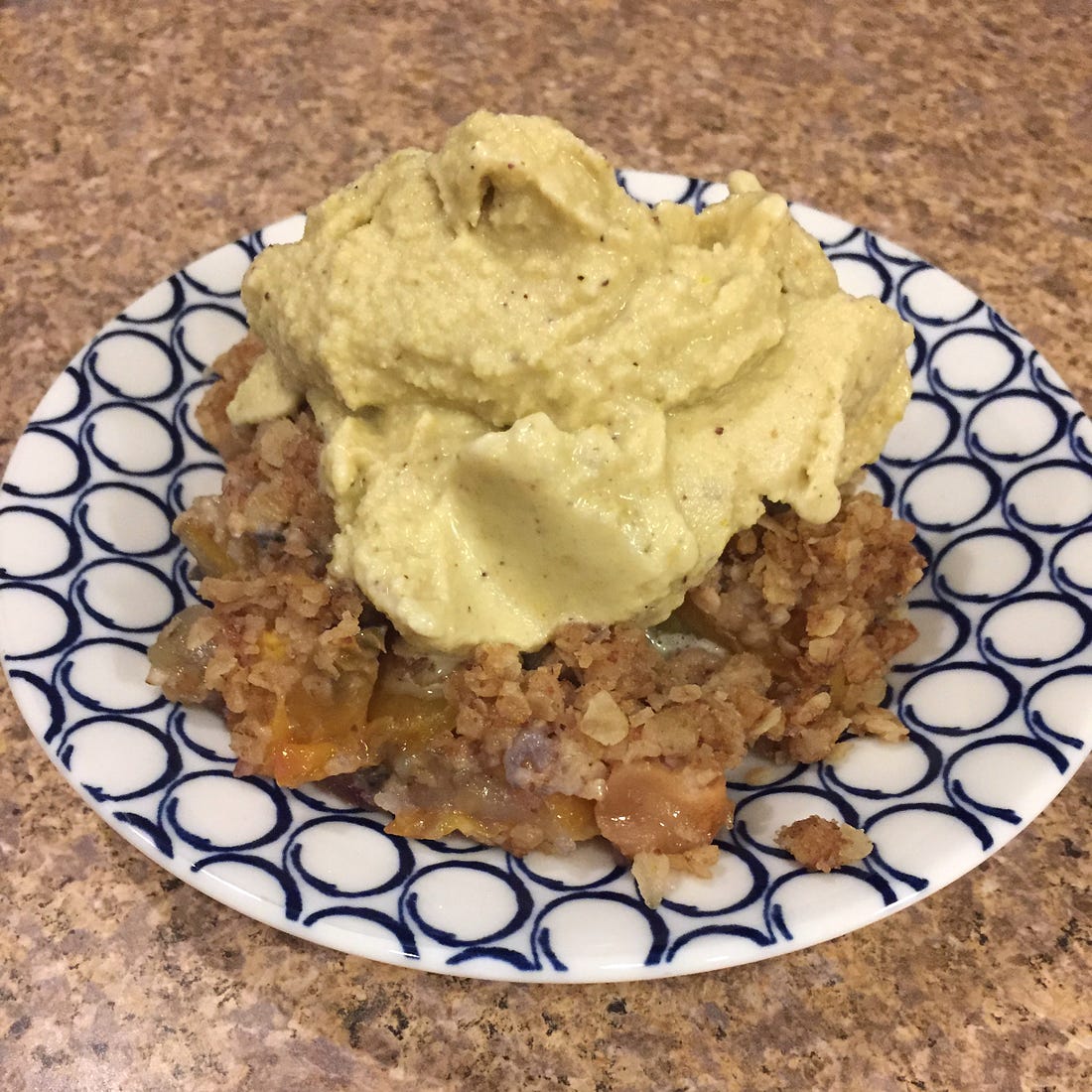 on a blue and white patterned plate, a mess of peach and apple cobbler with a soft-looking scoop of pistachio ice cream on top.