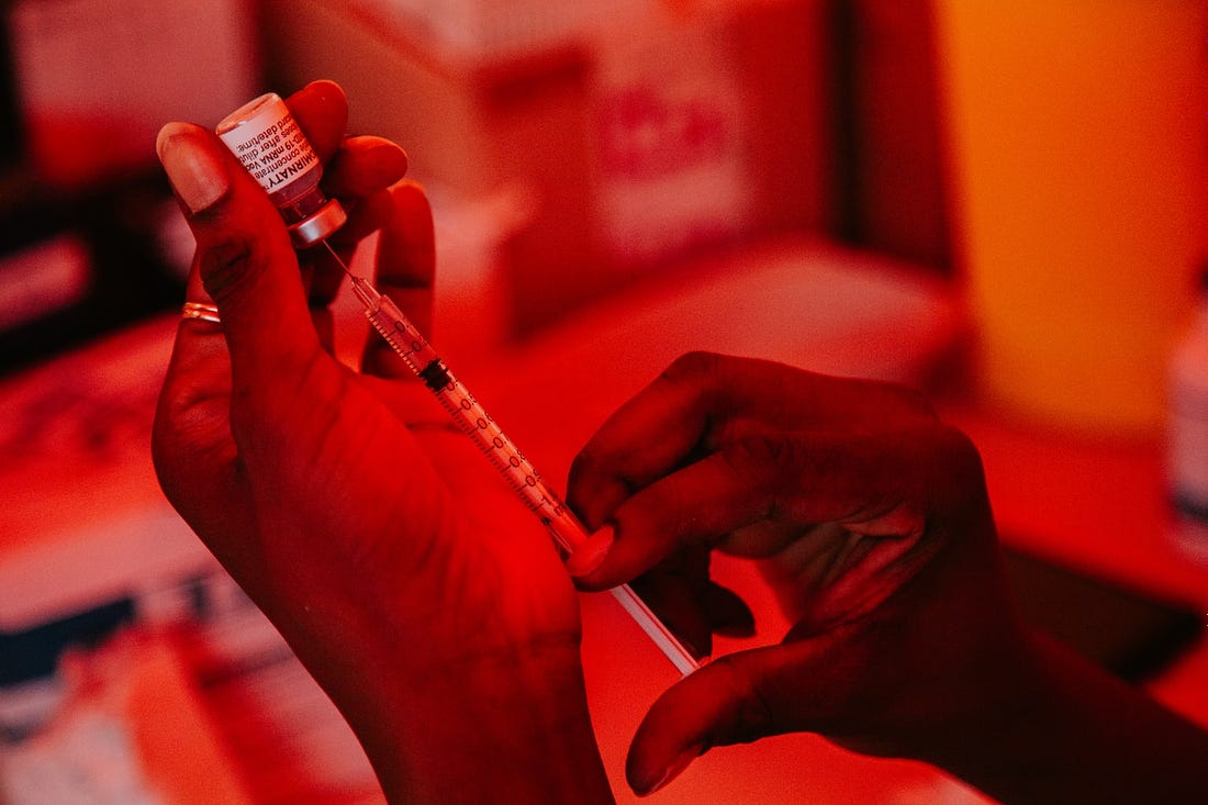in dark red lighting, a close-up of two Black hands filling a needle with the liquid vaccine
