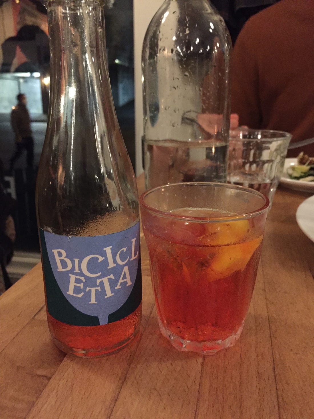 A restaurant table with a small glass full of a pinkish-red cocktail, with a slice of orange rind floating on top. Next to the glass is a small glass bottle with a blue label that reads 'bicicletta' in stylized capital letters. In the background, behind a tall glass bottle of water, Jeff's hand picks up a fork.