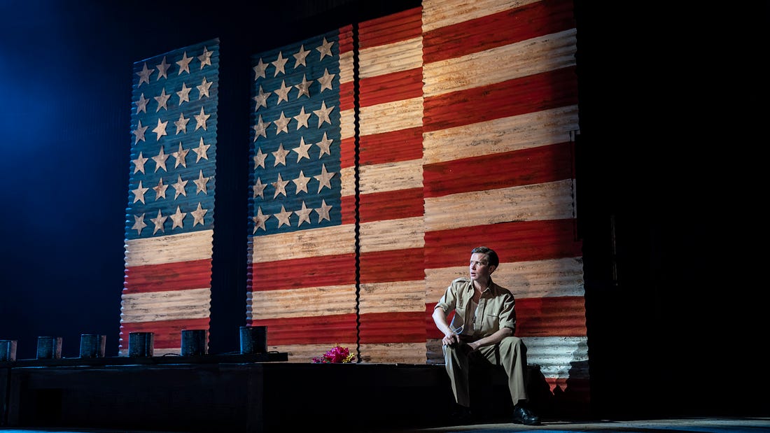 A young man wearing a 1940s US Army uniform sits in front of a fractured American flag.