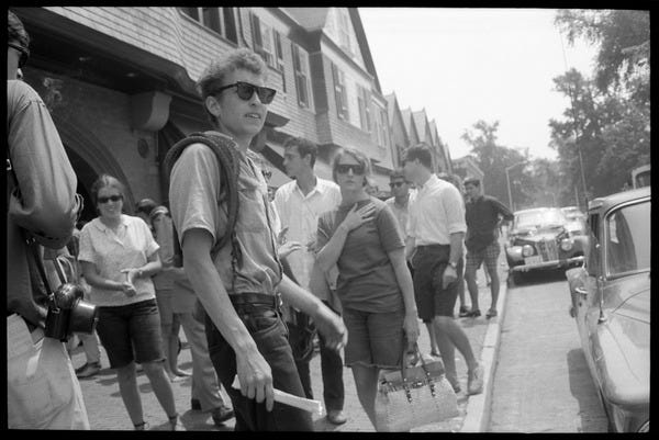 Bob Dylan, with bullwhip and sunglasses, walking on the sidewalk, Newport  Folk Festival: View from stage right, with stands in the background, July  27, 1963
