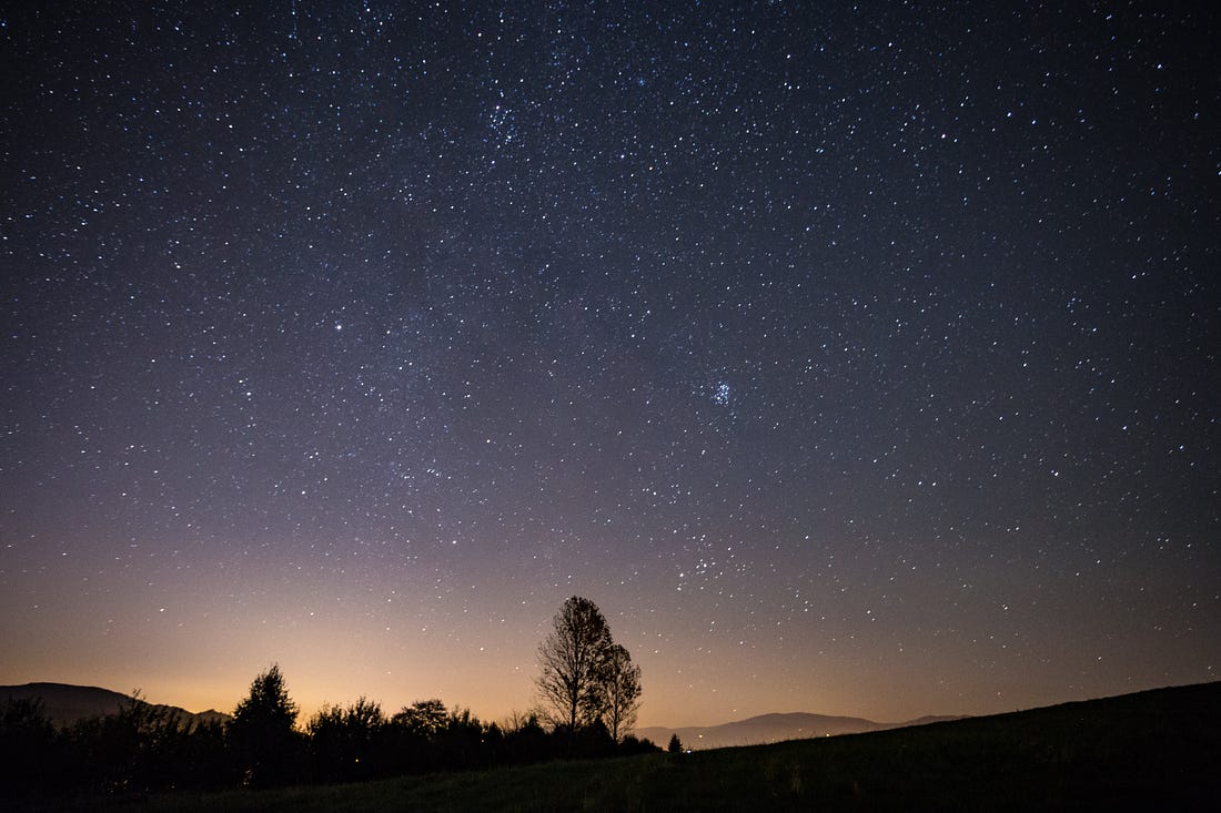 A sky full of stars, with a soft glow behind trees along the horizon line.