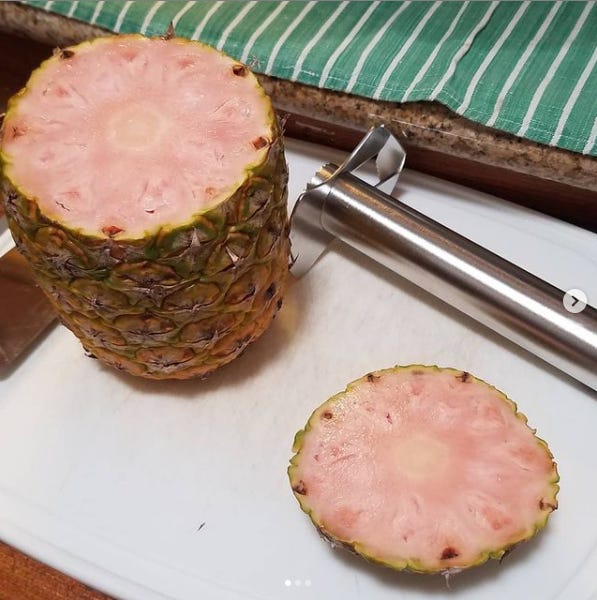 Whole pineapple with the top cut off standing on a cutting board next to a pineapple cutting gadget. The inner flesh of the pineapple is light pink.