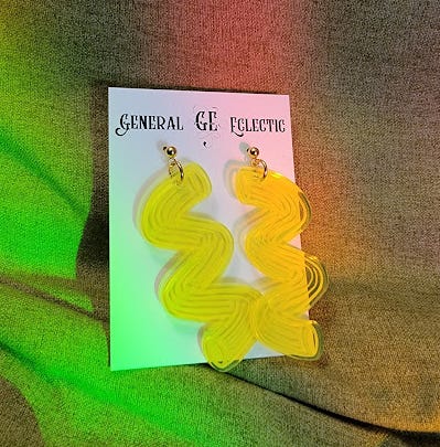 Photo of two bright yellow acrylic earrings shaped like cavatappi pasta. They are on a white cardboard back from General Eclectic Company. This sits on a green background. There are rainbows shining from prisms.