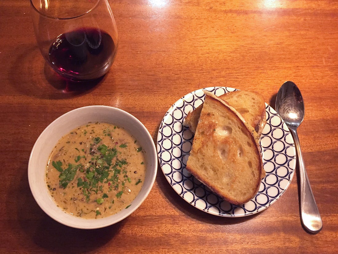 A small white bowl of creamy orangey mushroom soup topped with parsley and black pepper, next to a small plate with two halves of a grilled cheese stacked on top of each other. Above them and to the left is a stemless glass of red wine.
