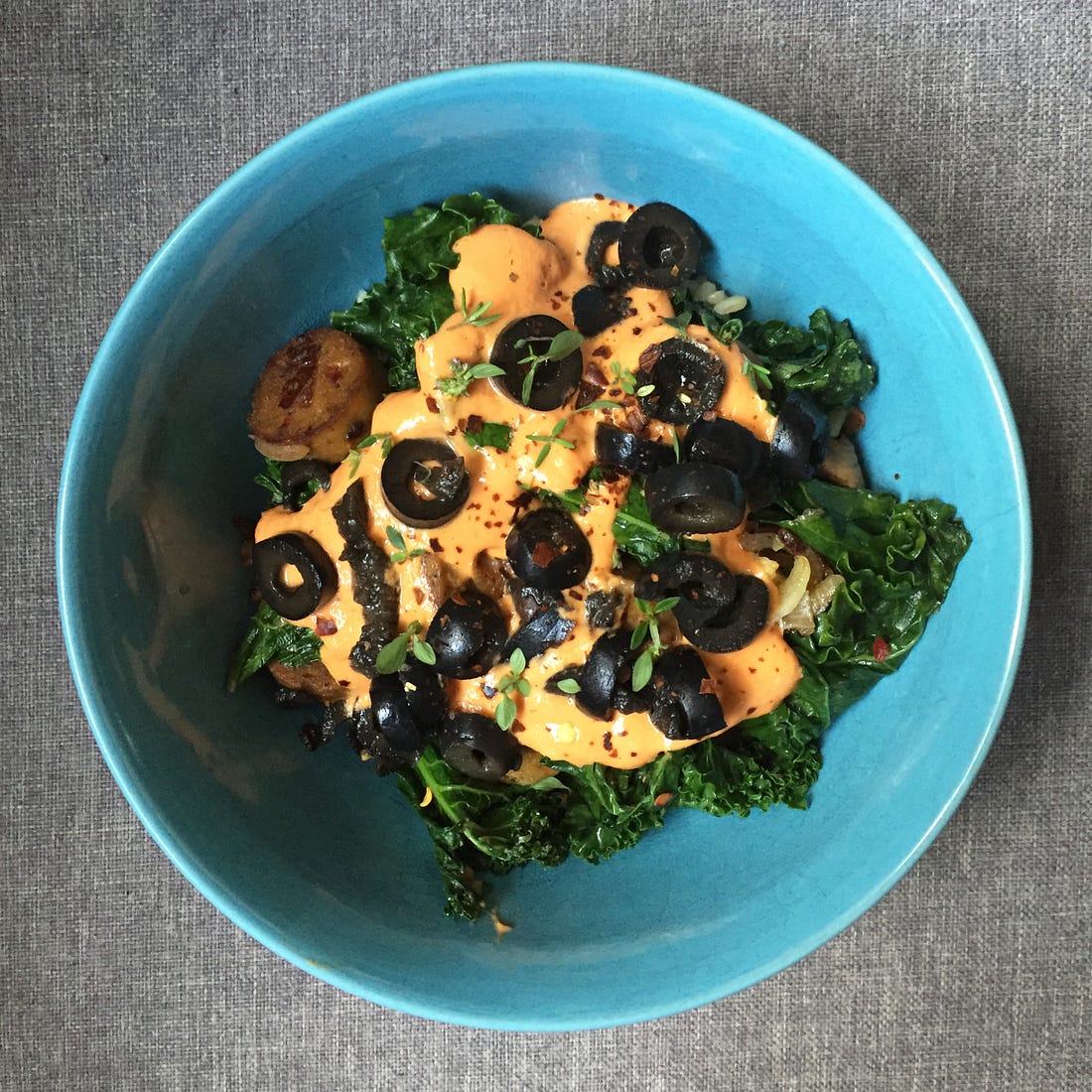 A blue bowl from above, filled with kale and sausage and covered in a cheesy sauce. Black olive slices, thyme leaves, and chili flakes are sprinkled on top.