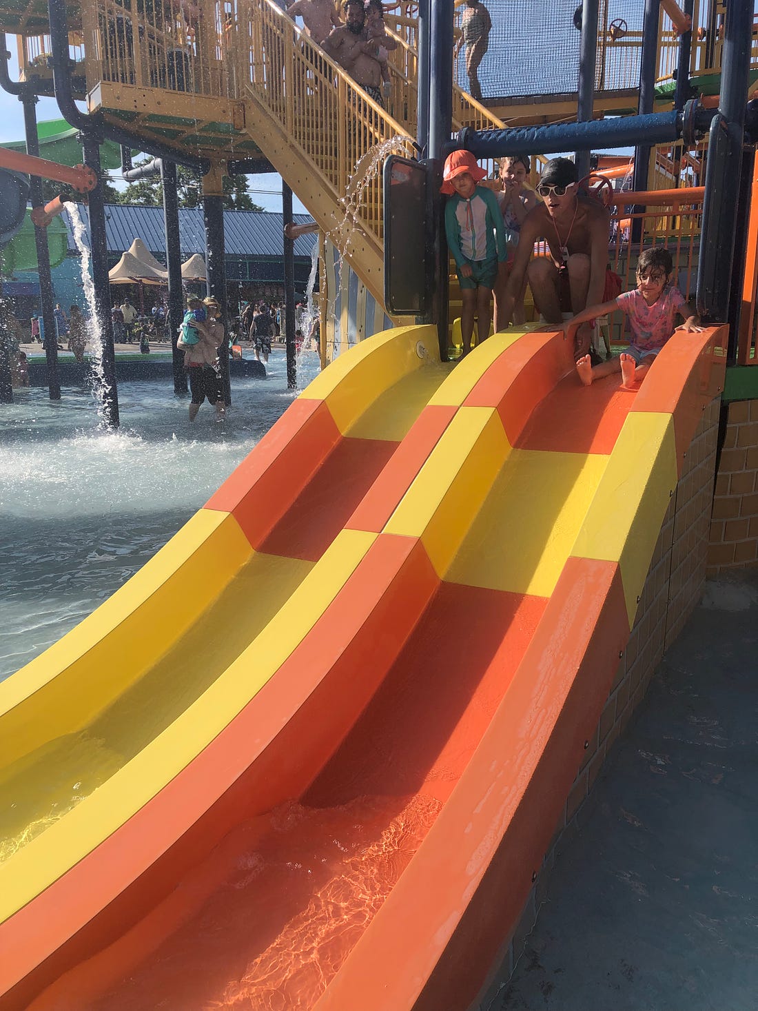 Photo shows a climbing structure with a small side-by-side water slide with two kids and a lifeguard.