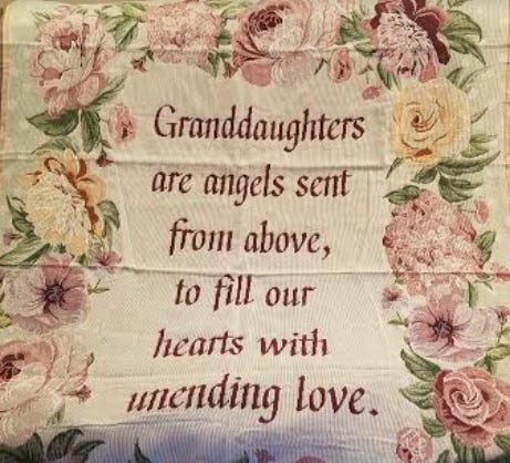 Photo of woven blanket. Primary color is cream and there are cream and white very large flowers around the border. In the center in dark pink words it says, “Granddaughters are angels sent from above, to fill our hearts with unending love.”