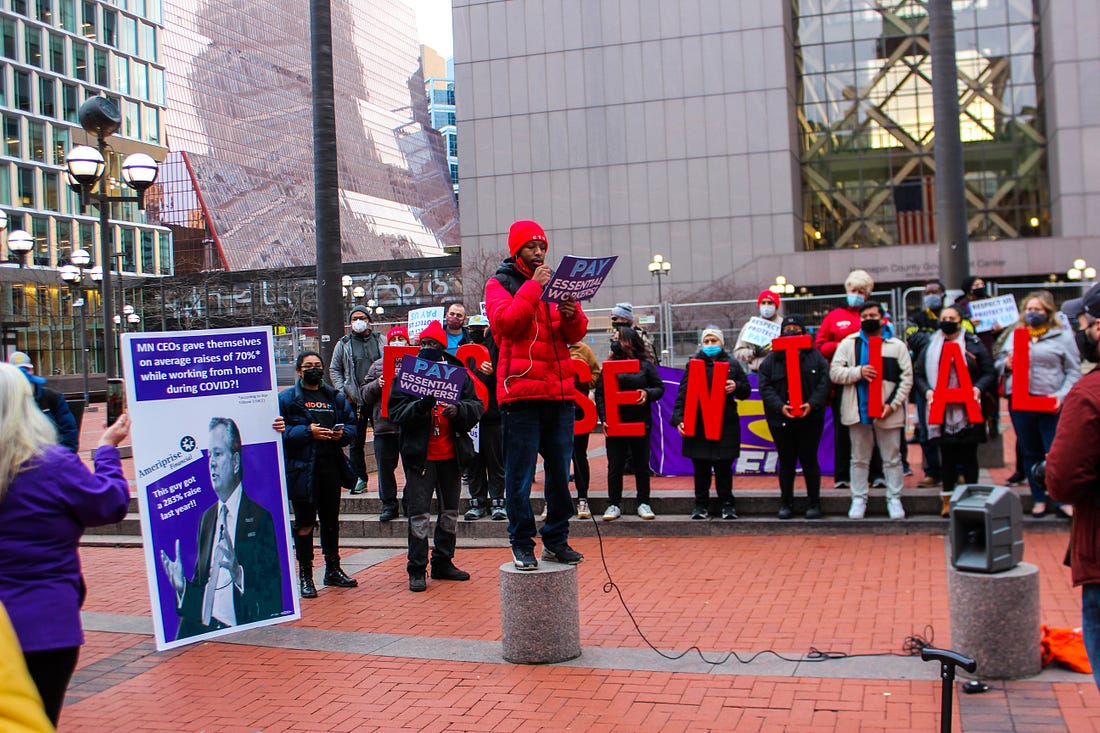 a line of people holding big red letters spelling "ESSENTIAL" stands behind a black man in a red coat and hat holding a "pay essential workers" sign