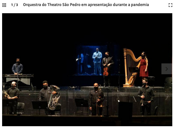 Imagem mostra palco de teatro com músicos de máscara em distanciamento sob fundo escuro.