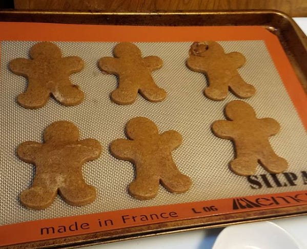 Six gingerbread cookies shaped like people laying on a silicone baking mat. The cookie on the upper right looks like its head exploded in the oven.