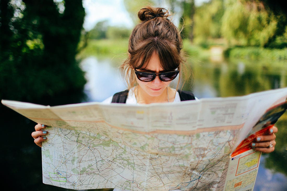 A woman standing outside with green trees and a lake in the background that is blurred, she is wearing sunglasses and she is holding a map open that spans across the entire bottom of the image.