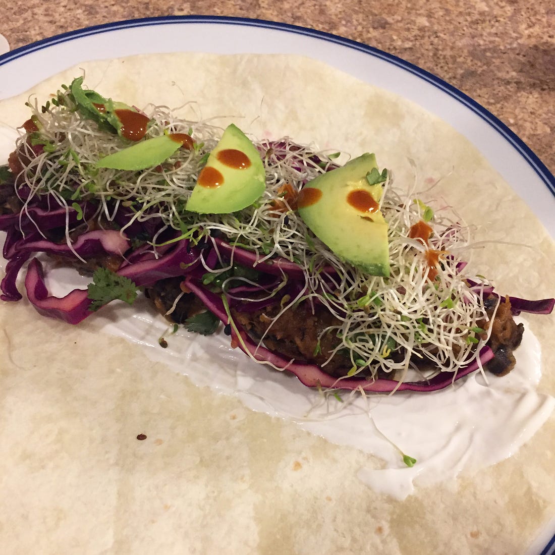 A burrito, prior to being rolled up, sits on a large round plate.. The bean and squash filling is just visible on top of a smear of sour cream and beneath red cabbage slaw, lots of sprouts, and slices of avocado dotted with hot sauce.