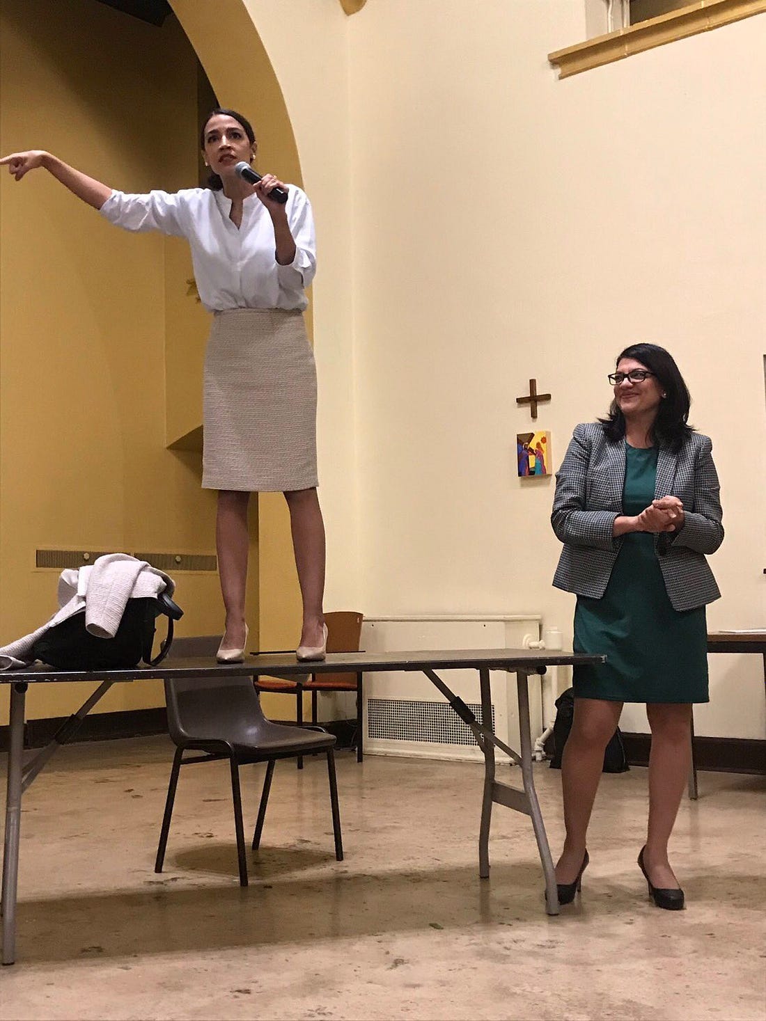 Rep. AOC stands on a table talking into a mic as Rep. Rashida Tlaib looks on smiling