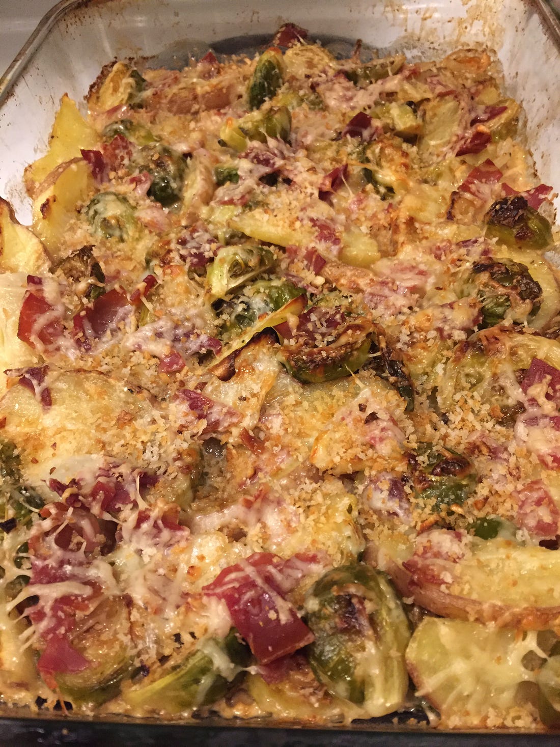 a baking dish full of the gratin described above. Breadcrumbs are scattered over the top, and beneath a thin layer of browned cheese are pieces of prosciutto, potato wedges, and halved brussels sprouts with onions throughout.
