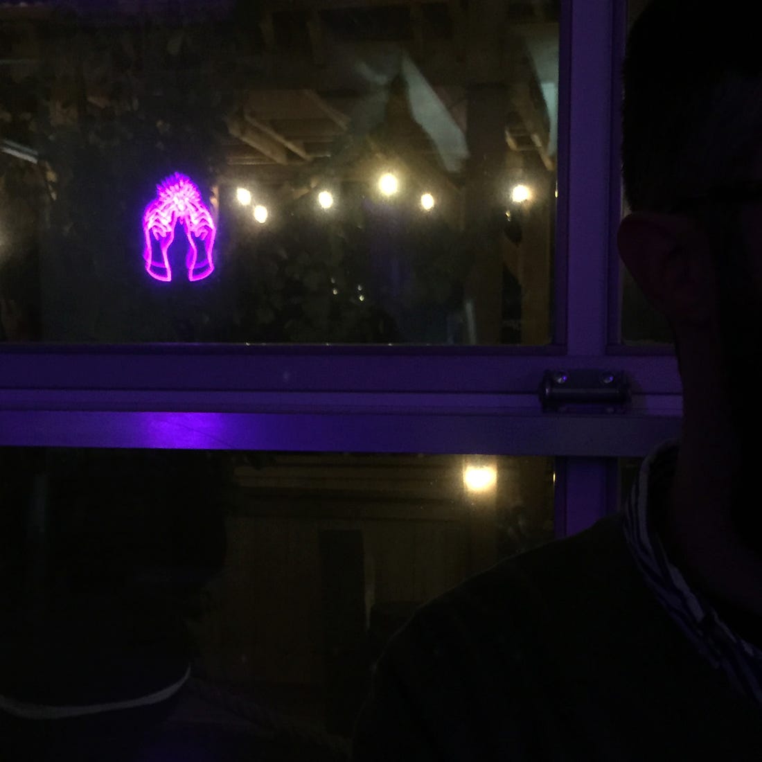 The pink neon logo of House of Funk (two hands with a grain levitating between them) reflected in a dark window. Other lights are reflected in the window, and a barstool is visible in the bottom corner of the photo.