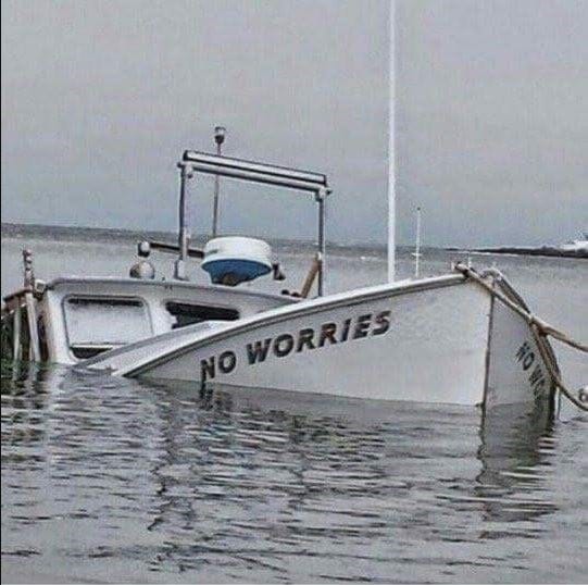 Photo of a white boat that is sinking. It is mostly above water. The front of the boat is still above water and it has the boat name on it. The boat is named "No Worries"