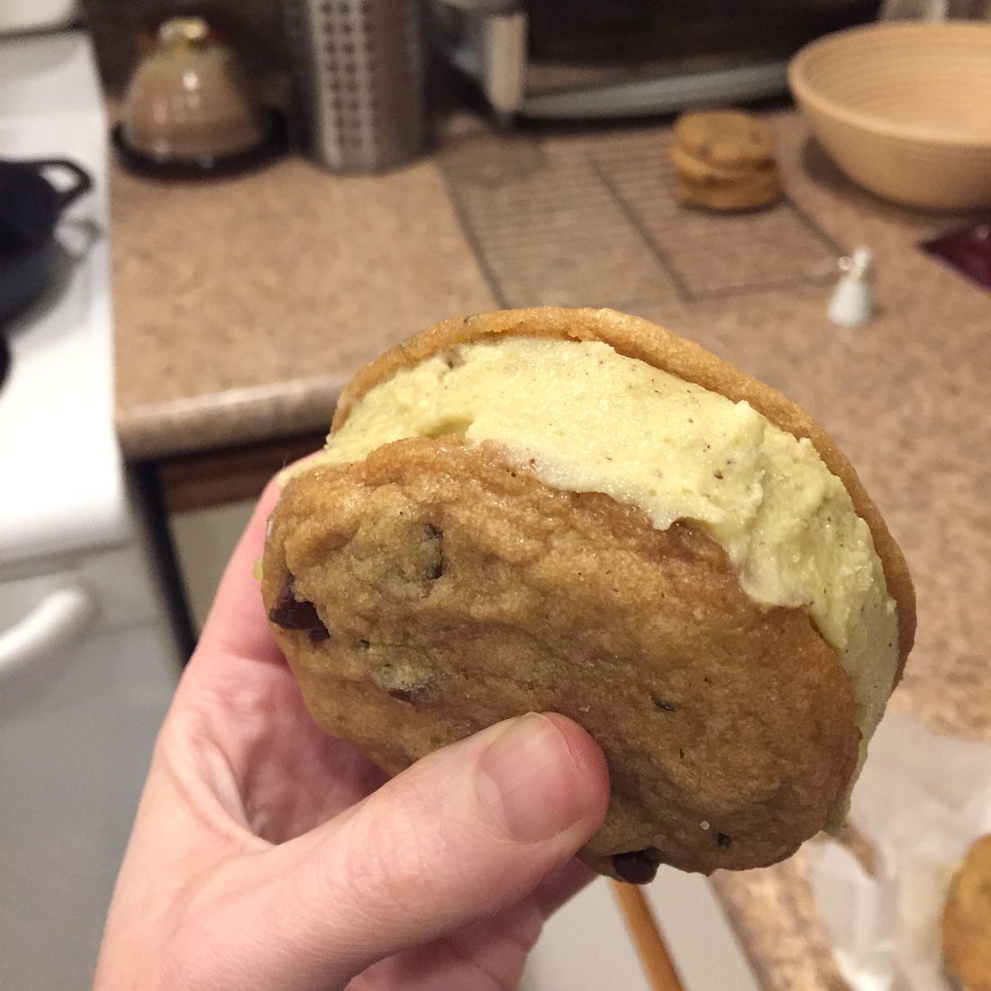 My hand holding the ice cream sandwich described above. Green pistachio ice cream is neatly spread between two of the cookies. In the background, a small stack of the cookies is visible on the counter next to the stove.