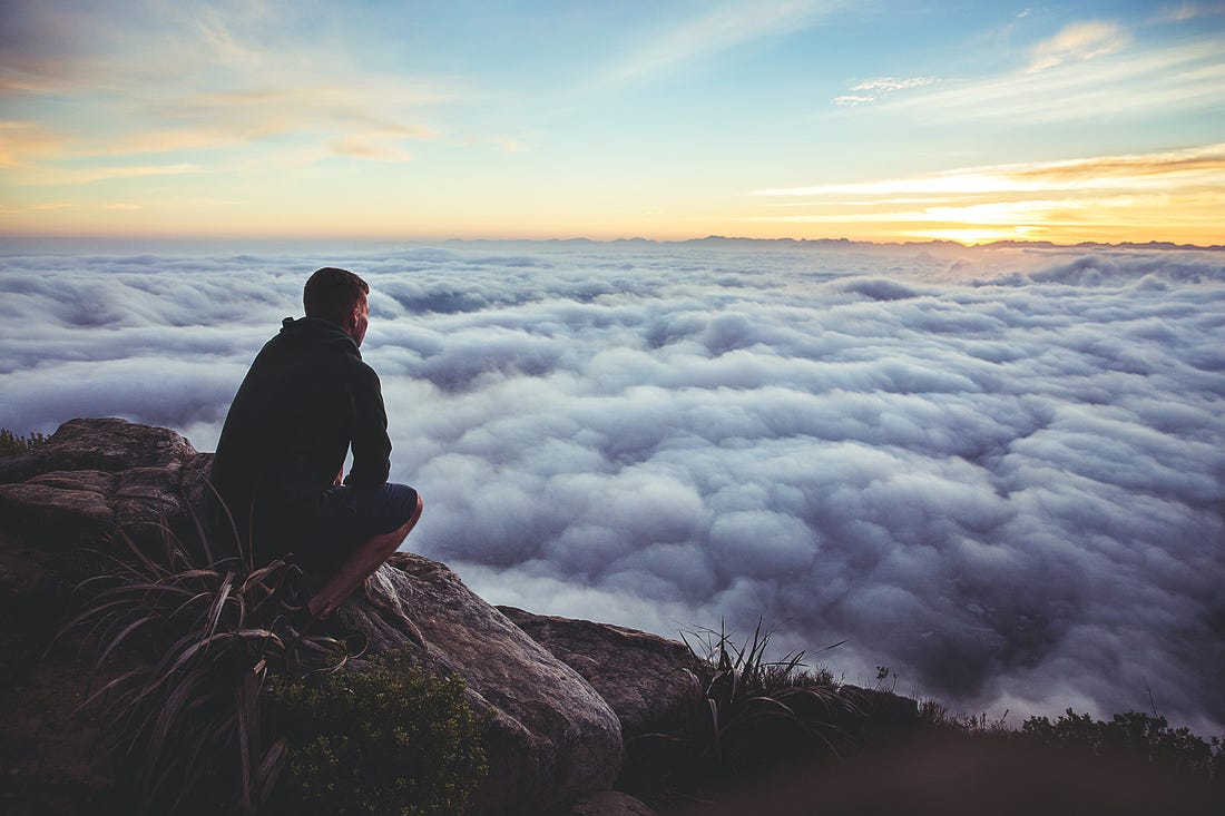 Man sitting on a mountain top and looking at the clouds below.