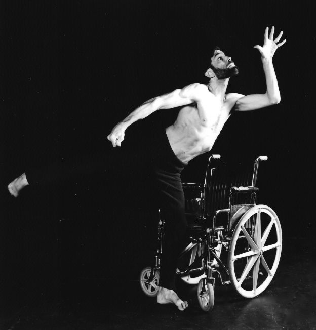 A black and white photograph shows a white man in the middle of a dance on stage. He is standing on one foot, kicking the other behind him, reaching one hand up in the direction of his gaze while his other arm parallels his other leg. Next to him is a manual wheelchair.