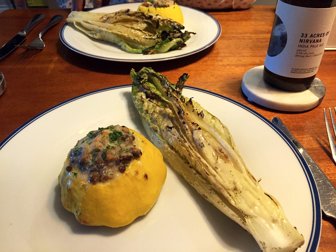 Two large white plates across from each other on a wooden table, each with a small patty pan squash stuffed with lentils and topped with cheese and parsley. Next to the squash is half a grilled romaine heart with Caesar dressing. On a coaster nearby is a bottle of beer.
