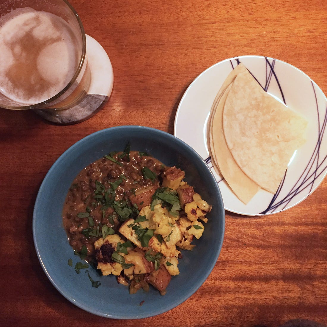 From above, a blue bowl with dal on one side and roasted aloo gobi on the other, sprinkled with cilantro. On a small plate are quarters of a tortilla, and on a coaster in the upper left is a glass of IPA.