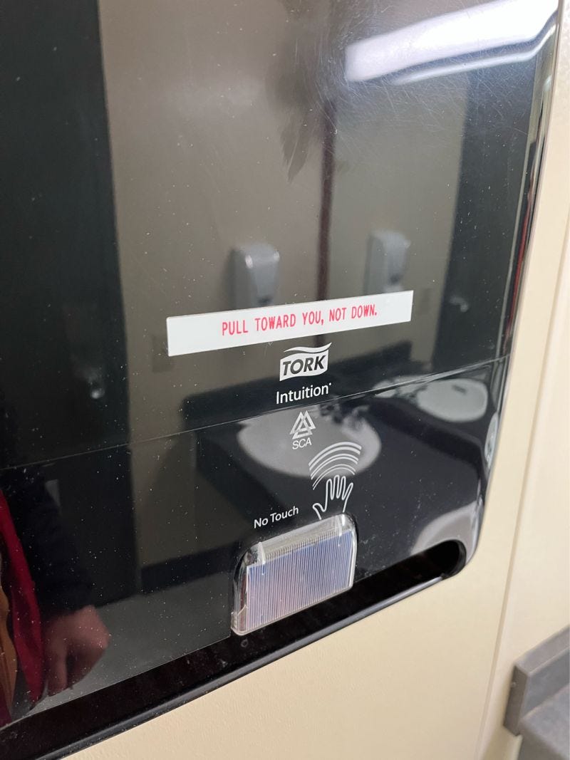 Paper towel dispenser with a sticker saying "Pull toward you, not down" indicating a design issue.