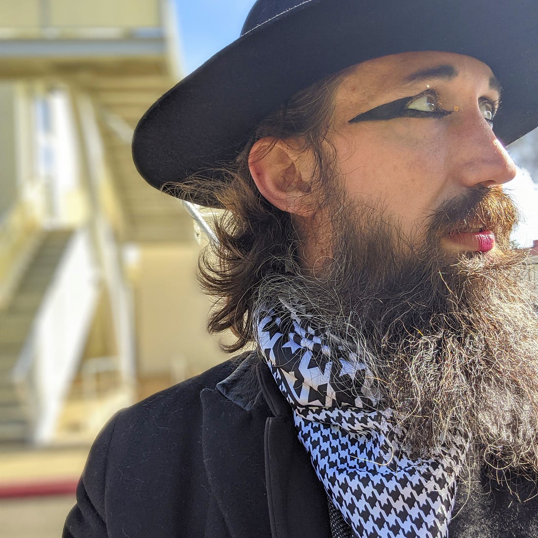 Jason Wyman looking to the left wearing a black wide-brimmed hat, a houndstooth silk scarf, and a black wool coat. Jason has long brown hair and a big bushy brown beard. They are wearing pink lipstick and thick black pointed eyeliner. Jason is set against a pale yellow home and a pale blue sky, both of which are blurry. 