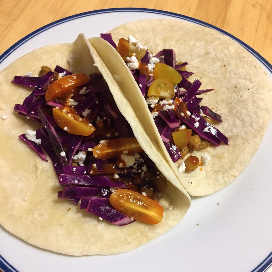 On a white plate, two flour tortillas filled with the taco filling described above. Most visible is the red cabbage slaw, crumbles of feta, and pieces of tomato with hot sauce overtop.