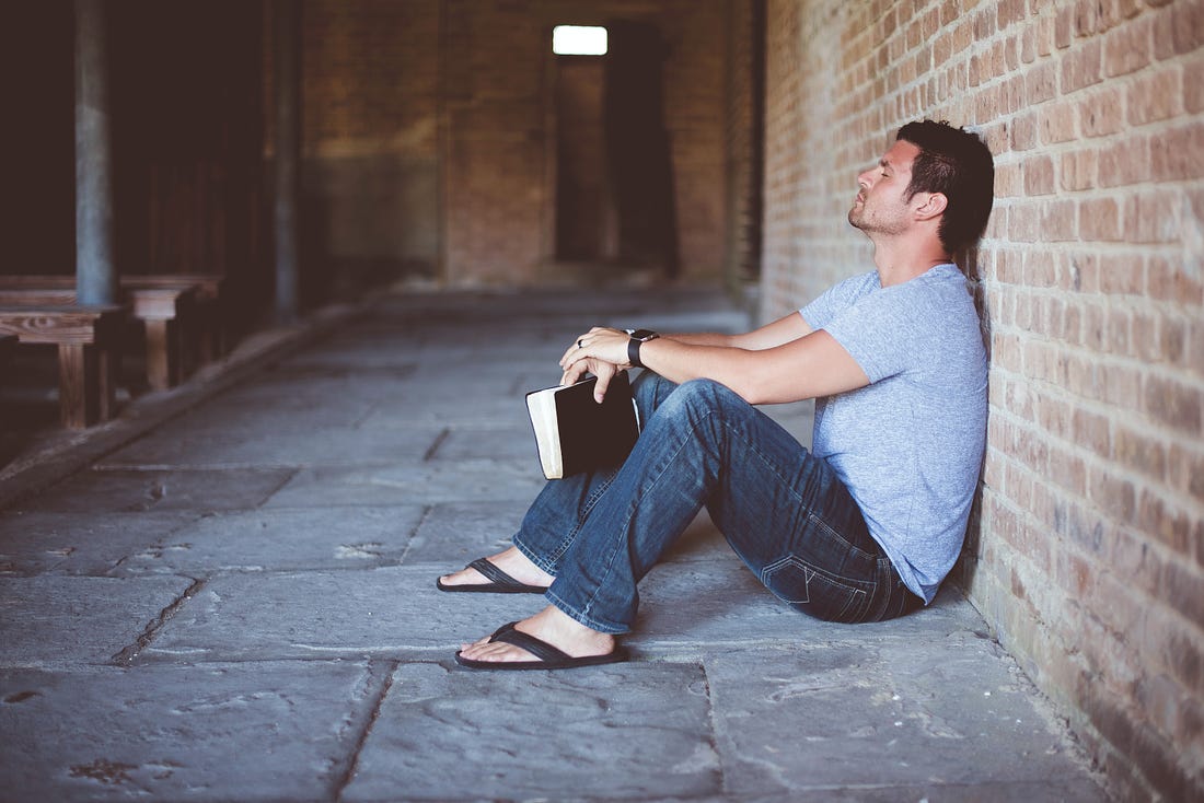Man sitting on the floor with his back against the wall and a Bible in his hands in contemplation