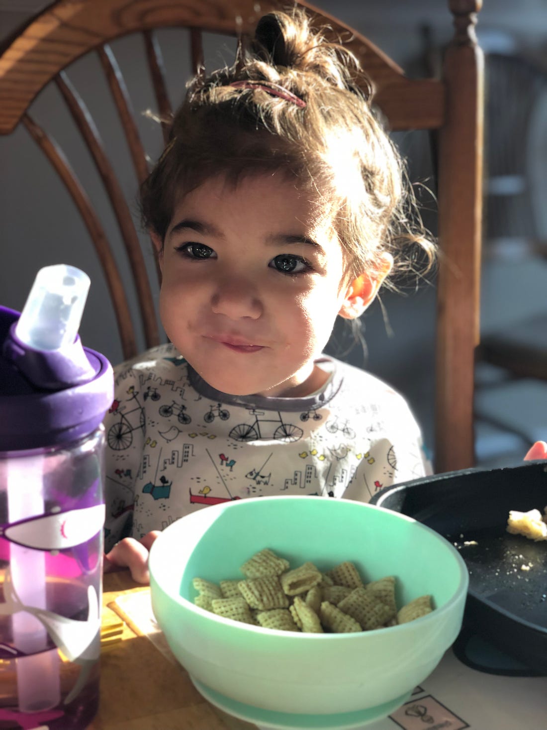 Photograph of a brown haired toddler sitting in a chair with a bowl of cereal in front of her.