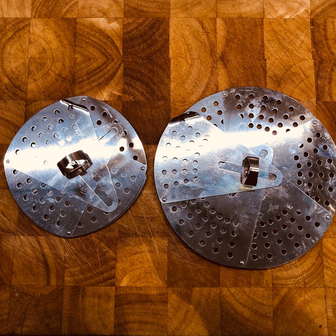Two metal adjustable drop lids. The lid on the left is smaller in diameter. The lid on the right has been expanded so it is larger. 