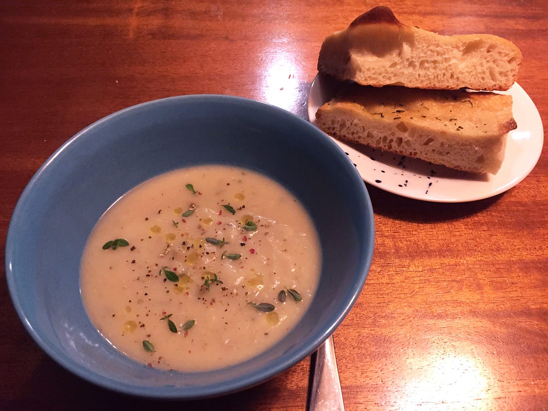 a blue bowl of potato soup with leaves of thyme, ground pepper, and a drizzle of oil on top. To its right is a small plate with two piece of focaccia bread. The sliced edge shows lots of holes, and the top of the bread is lightly browned, with a dusting of rosemary.