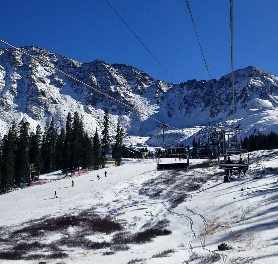 a craggy mountain beneath a clear blue sky. Cables zip overhead. Snowboarders and Skier enjoy an early-season run below