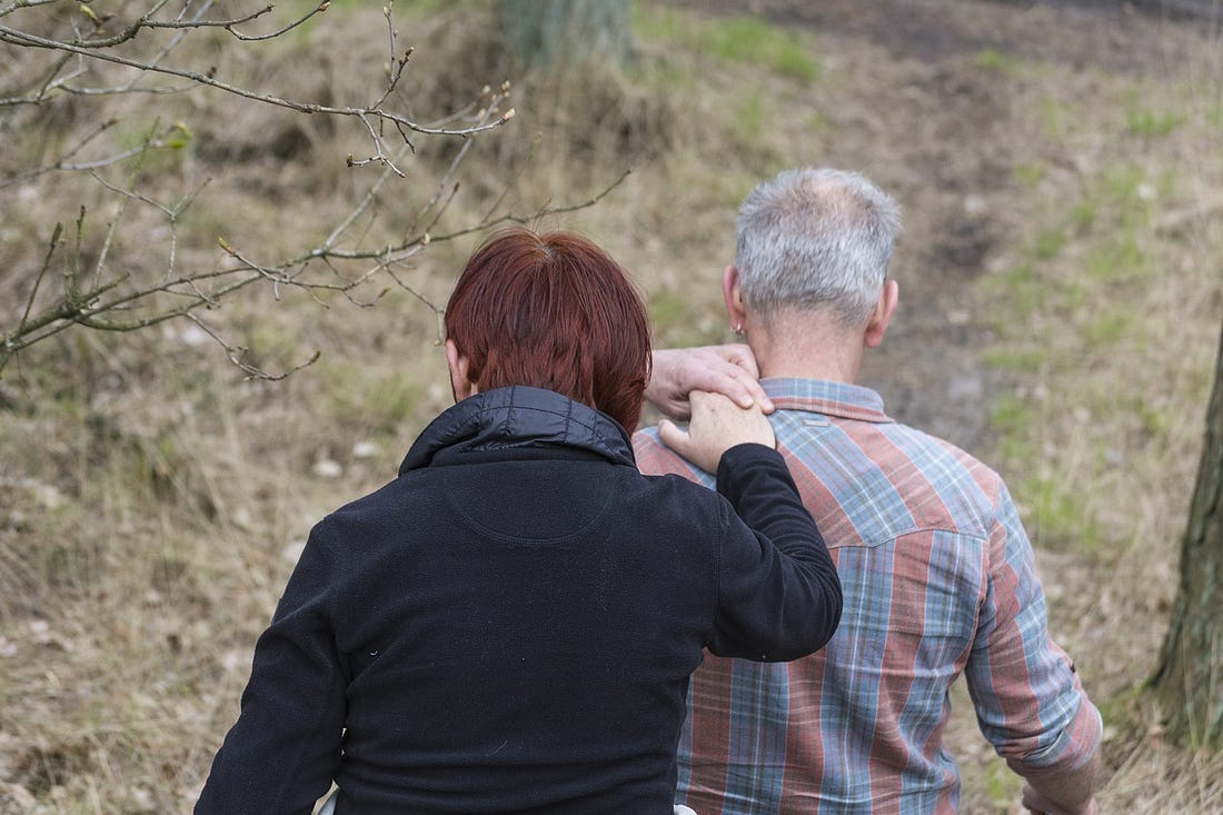 A man leads a woman along a path near a tree by clasping her hand on his shoulder.