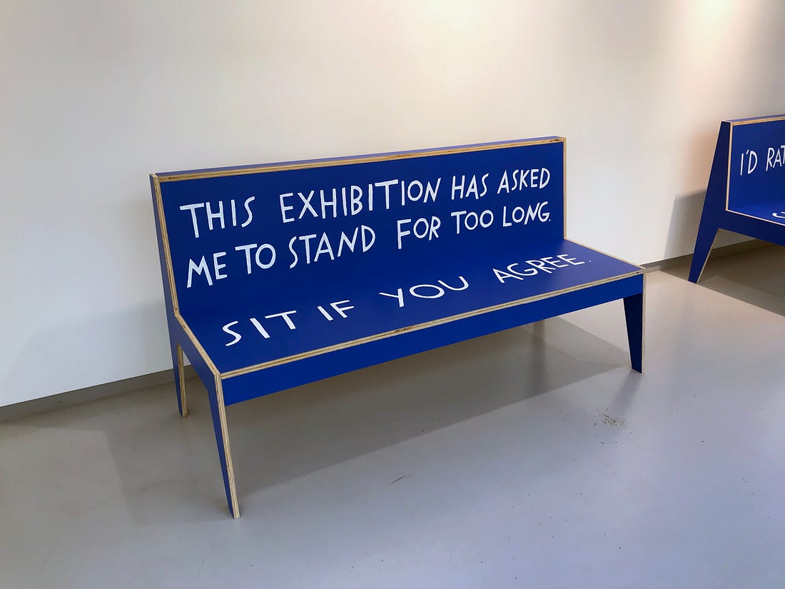 A blue bench with hand-painted text that reads, “This exhibition has asked me to stand for too long. Sit if you agree.”