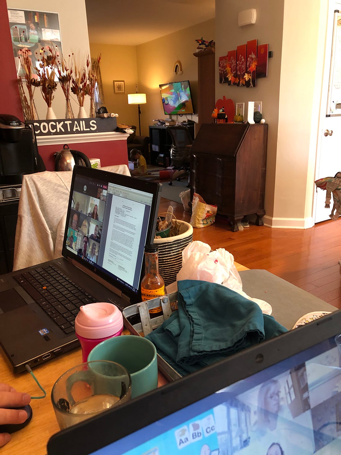 Photo shows a toddler in the background with the television on, two laptops at a dining room table with an IEP meeting in session. Photo shows a toddler in the background with the television on, two laptops at a dining room table with an IEP meeting in session.