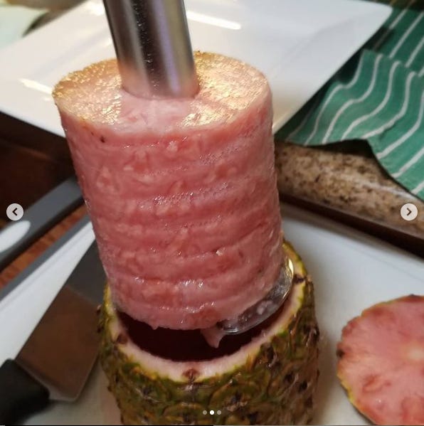 The insides of a pink pineapple are being lifted out in a complete spiral on a stainless steel pineapple cutting gadget