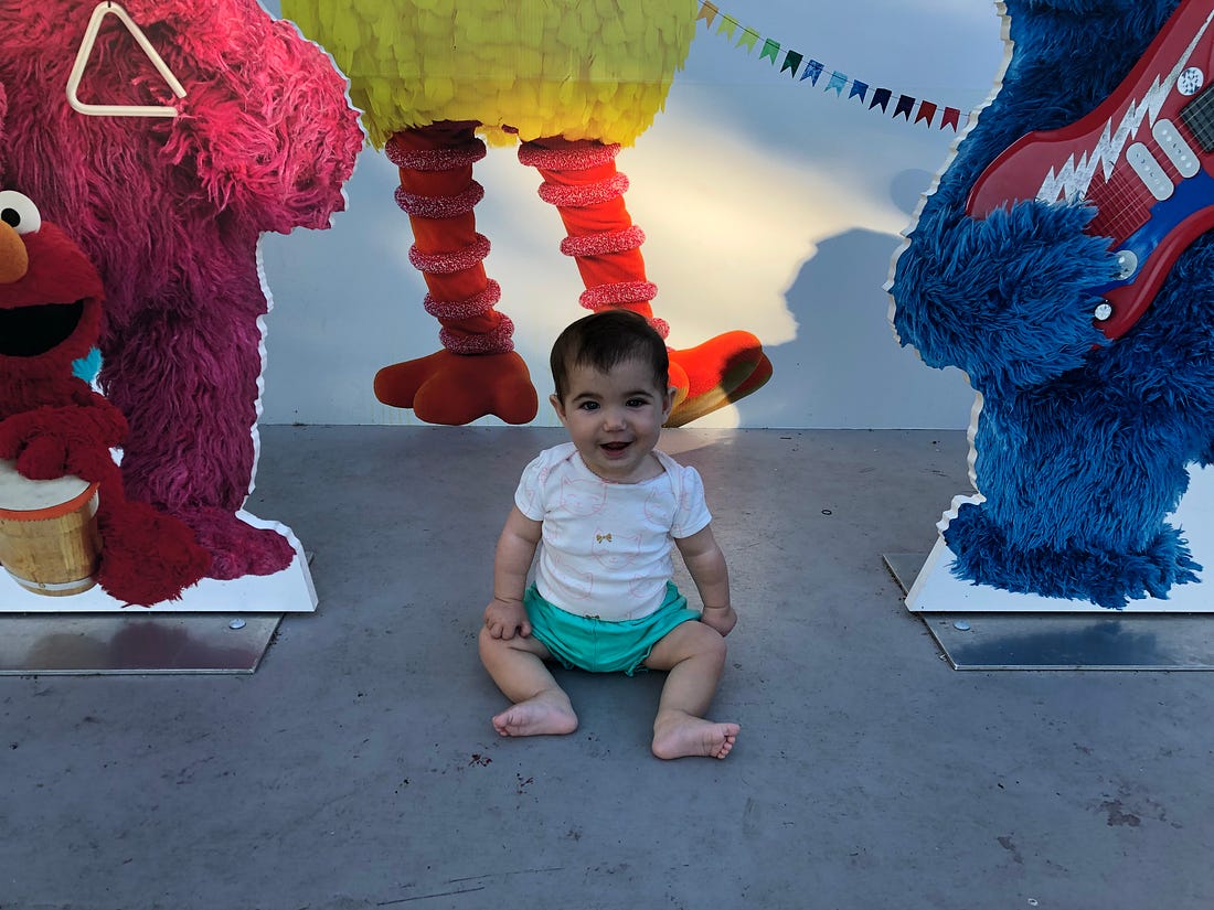 Small baby with a giant smile sits upright between cutout figurines of sesame street characters.