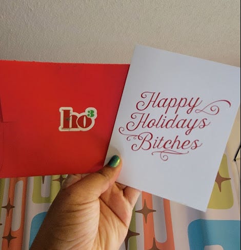 Photo of Patricia’s hand holding up a red envelope and a white card. On the envelope is a sticker that has the word, “ho” to the third power (ho cubed). The card has red script that says, “Happy Holidays Bitches”
