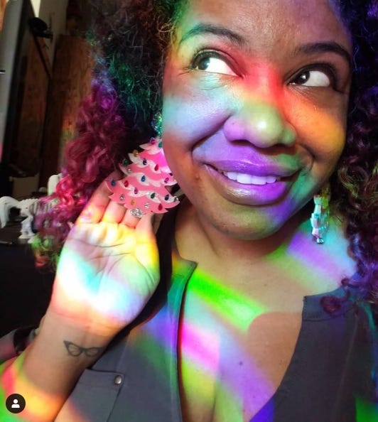 Photo of Patricia wearing large earrings that are acrylic pink Christmas trees with lots of glitter. She is covered in rainbows made by suncatchers in her window.