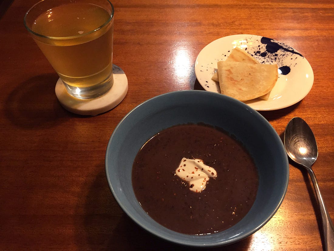 In a blue bowl, a puréed black bean soup. On top is a dusting of chili flakes, and a blob of crema in the centre. Above it to the right is a small plate with two wedges of quesadilla. Above it and to the left is a glass of lager.