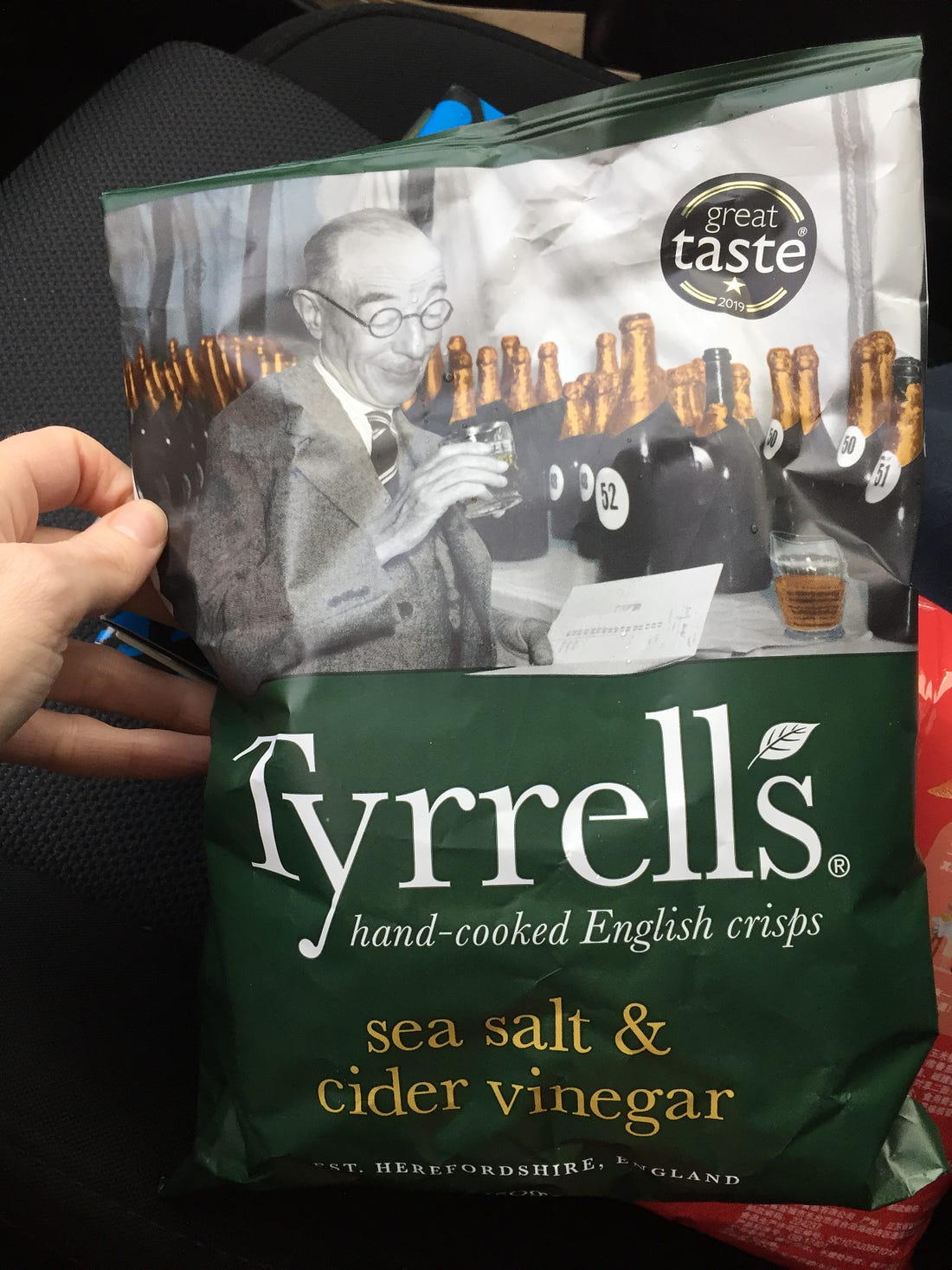 My hand holding up a bag of Tyrell's sea salt & cider vinegar chips. The bag is green with white and yellow text, and at the top is a black & white photograph of a man staring into a small glass of beer, with many more bottles in the background.