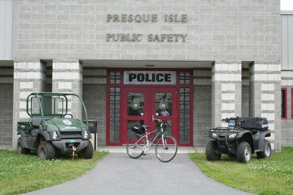 A golf cart, a bicycle and an all-terrain vehicle are parked in a row in front of a building. The building is gray concrete with four columns and a bright red entrance with double doors. Above the doors, are signs reading "Presque Isle Public Safety" and "POLICE".